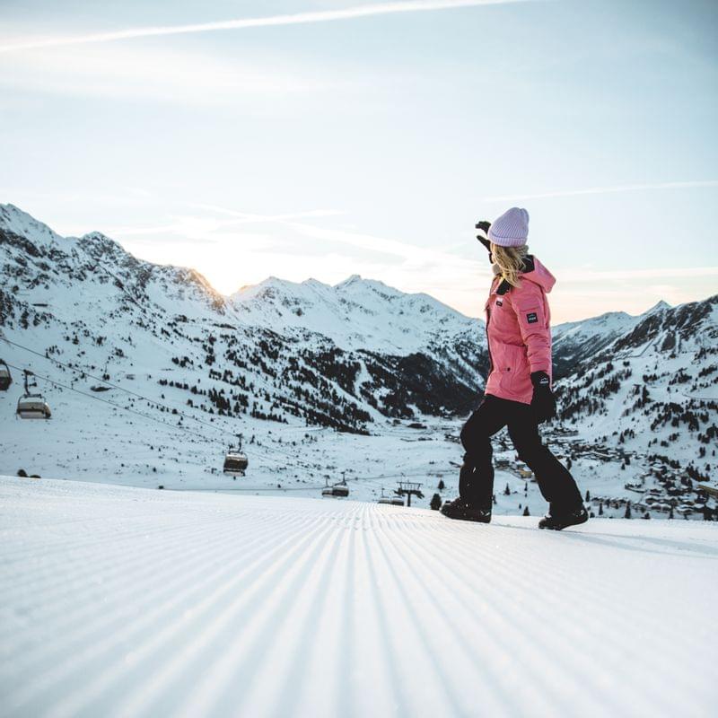 Frau in pinker Skijacke geht bei Sonnenaufgang über präparierte Piste mit verschneiten Bergen im Hintergrund